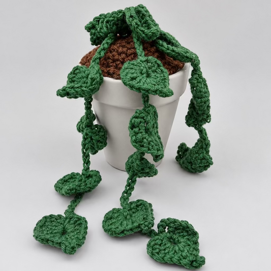 A green crocheted hanging leaf vine decoration draped over a small white pot filled with brown soil, photographed on a plain light background.