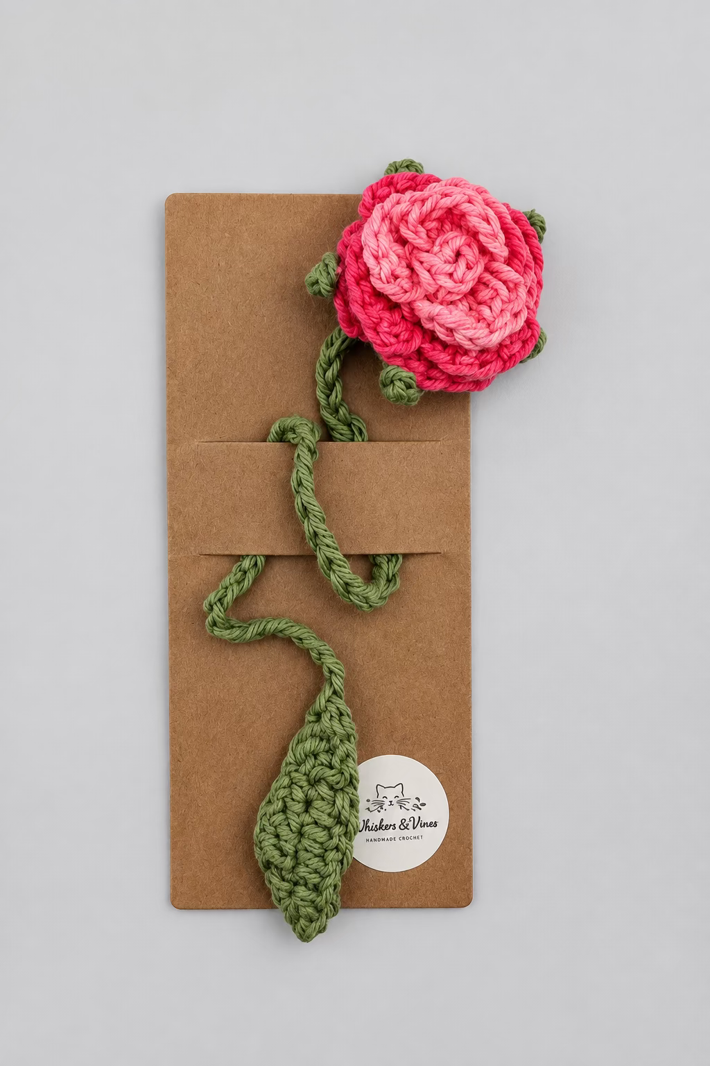 Crocheted pink flower and green stem on a brown card with a white background